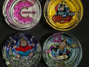 Cuatro ceniceros de cristal decorados con ilustraciones coloridas de la marca King Monkey. Uno tiene diseño de donut rosa con la palabra ‘Munchies’, otro es amarillo con un mono astronauta, y los otros dos muestran monos en diferentes poses y escenarios.