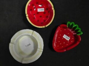 Tres ceniceros con diseños frutales y etiquetas de precio de 7,95 €. Uno tiene forma de fresa, otro simula una rodaja de sandía y el tercero es blanco con forma clásica redonda.